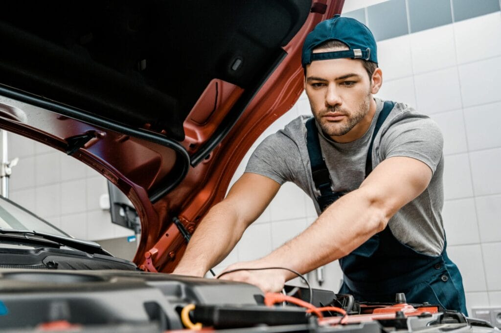 Mechanic in a cap and overalls working under the hood of a car in a garage.