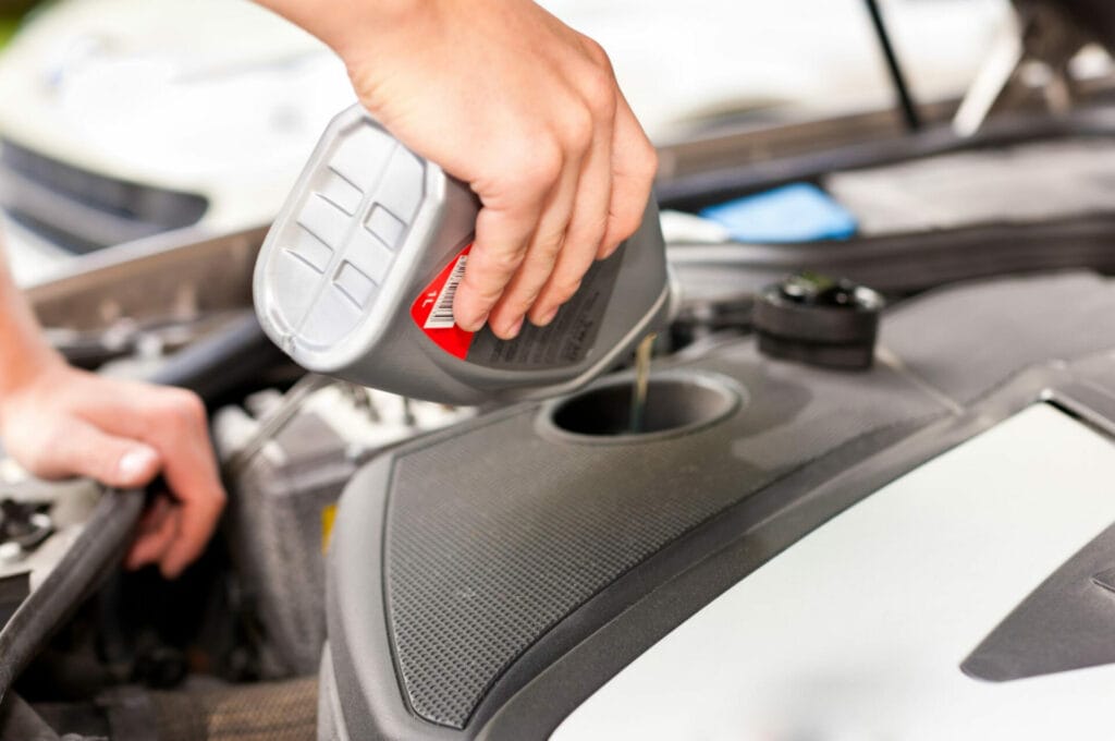 A person is pouring motor oil into an engine's oil fill hole under the hood of a vehicle.