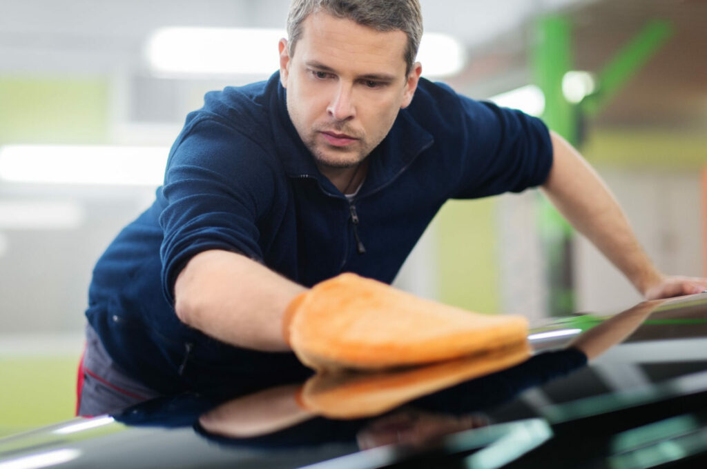 A person in a dark blue shirt uses an orange cloth to polish the surface of a black car.